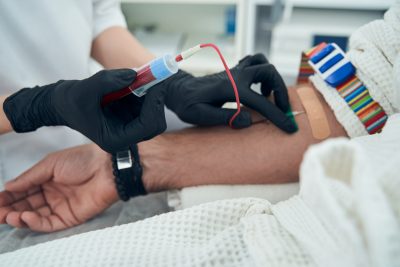 Close up of doctor's hands in black gloves holding test tube with blood while inserting needle into man's arm